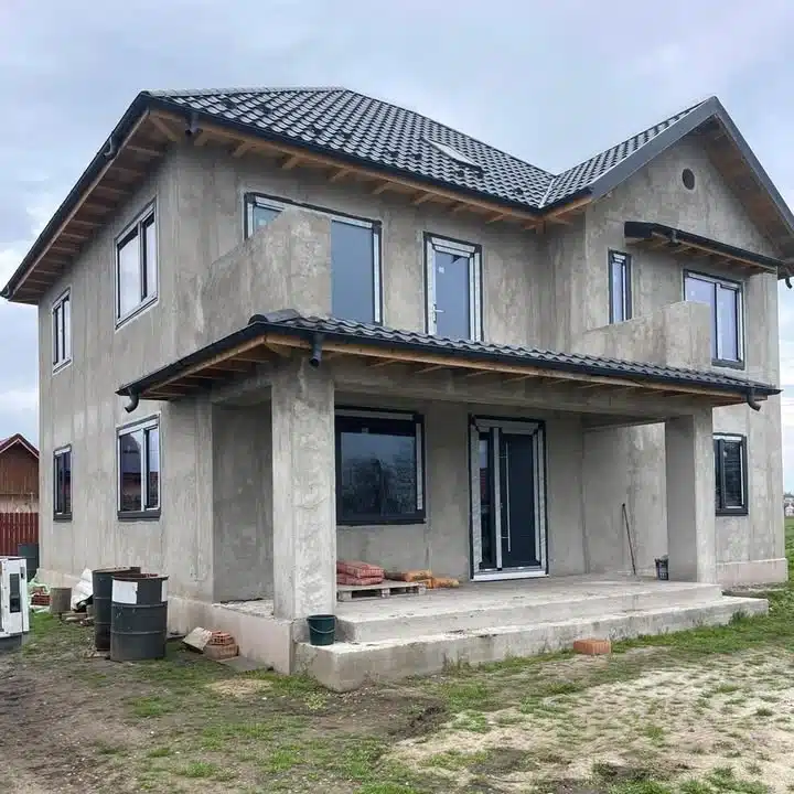 Casă în construcție cu tencuială gri și acoperiș negru.